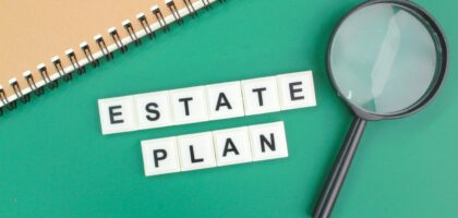 Estate Plan spelled with scrabble tiles on green background with notebook and magnifying glass.