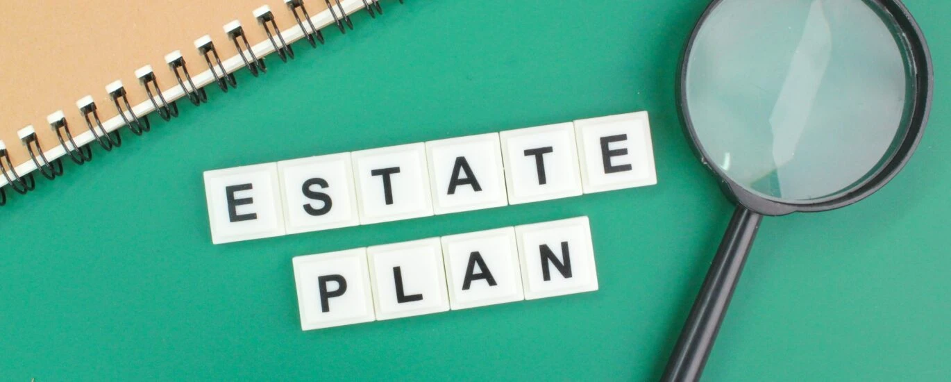 Estate Plan spelled with scrabble tiles on green background with notebook and magnifying glass.