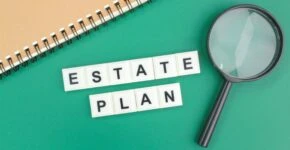 Estate Plan spelled with scrabble tiles on green background with notebook and magnifying glass.