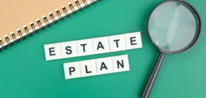 Estate Plan spelled with scrabble tiles on green background with notebook and magnifying glass.