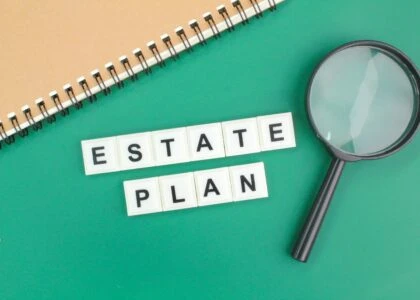 Estate Plan spelled with scrabble tiles on green background with notebook and magnifying glass.