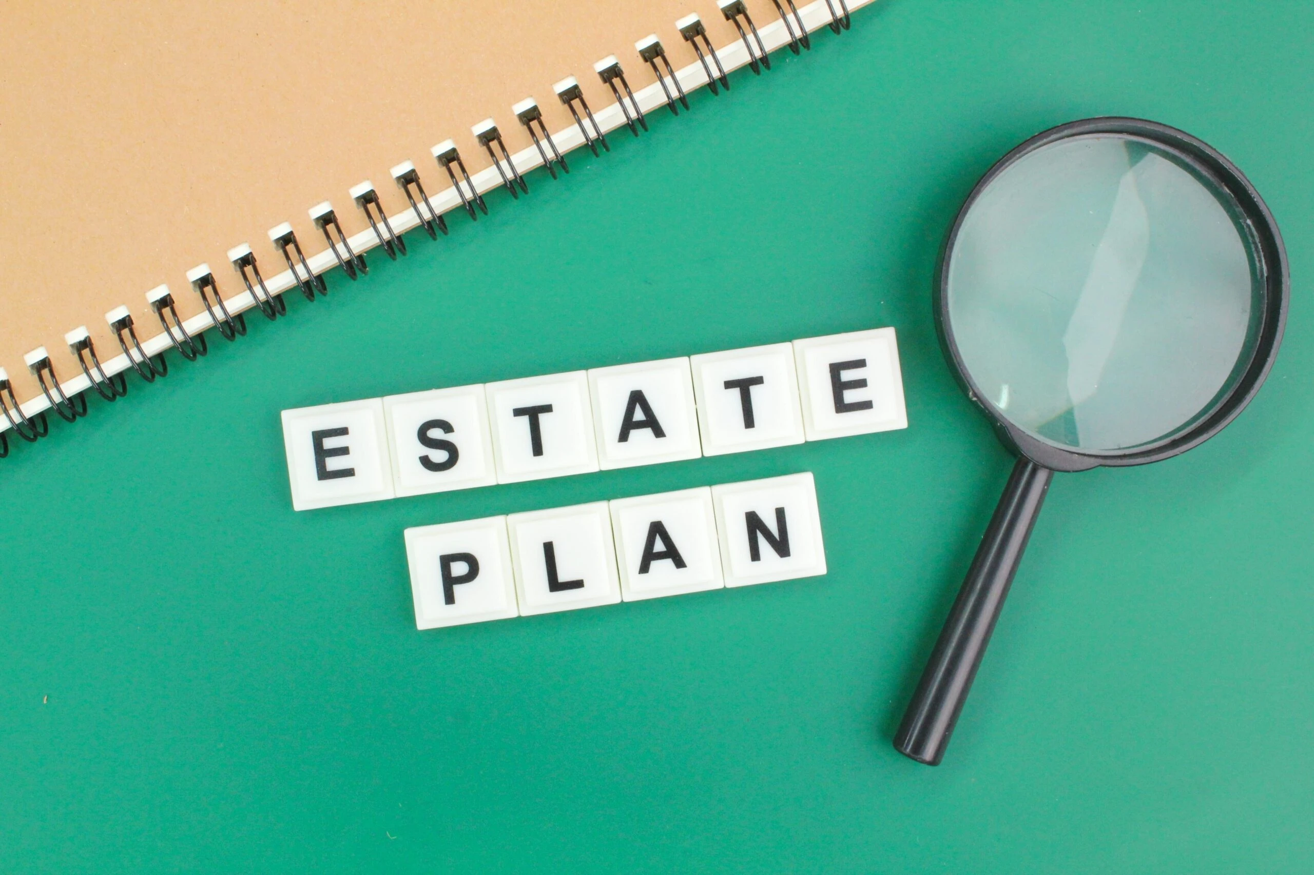 Estate Plan spelled with scrabble tiles on green background with notebook and magnifying glass.